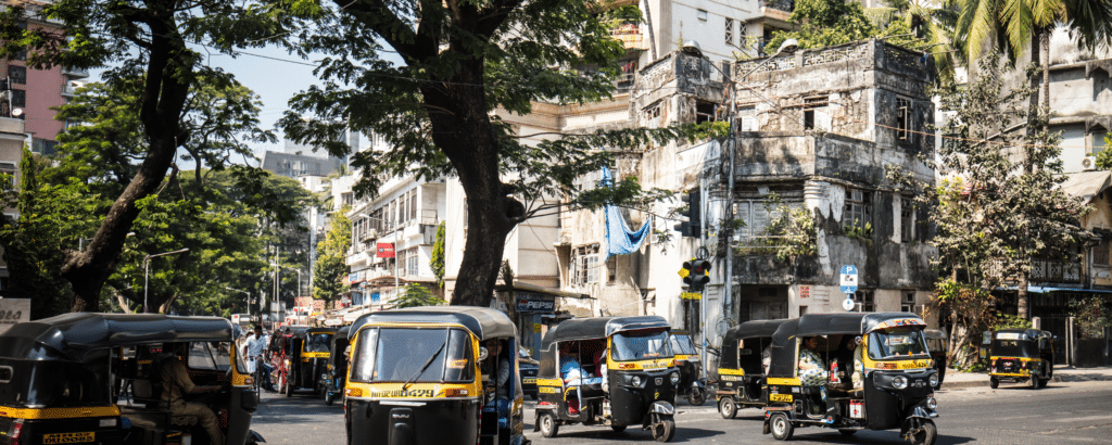 many auto rickshaws with passengers on the road. one old building. Two tall trees in Ghatkopar Khau galli.