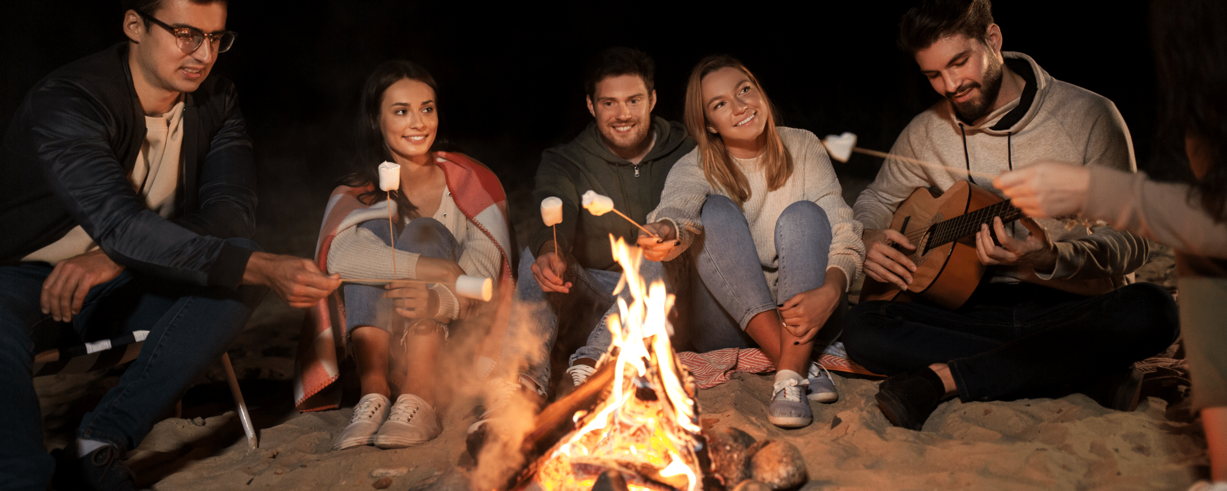 a group of youngsters enjoying bonfire and roasting marshmallows at the beach