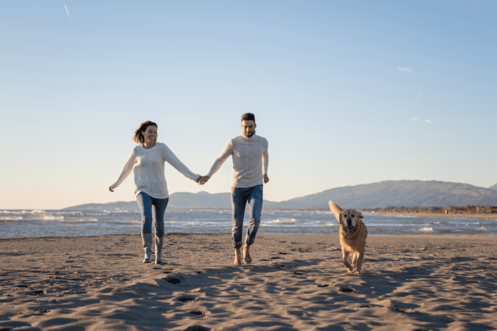 a man and woman holding hands and a dog all running on the rosie's dog beach