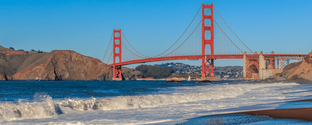 the golden gate bridge at baker beach. It is a red colored bridge with beach waves below.